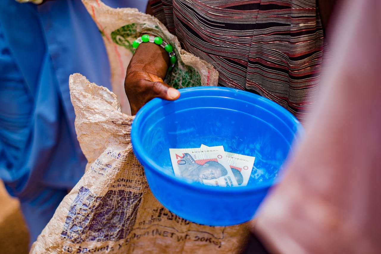 heros-img A close-up of a hand holding a blue bowl containing currency notes outdoors.