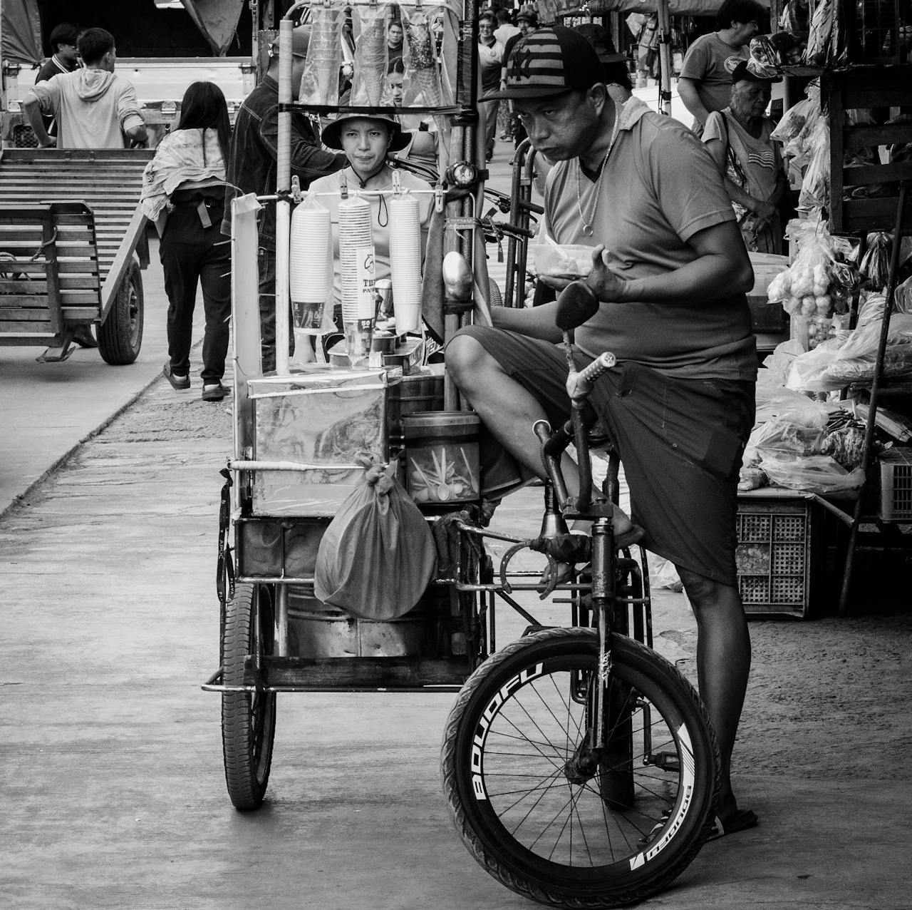 Black and white image of a street vendor with a bicycle cart in a bustling market.