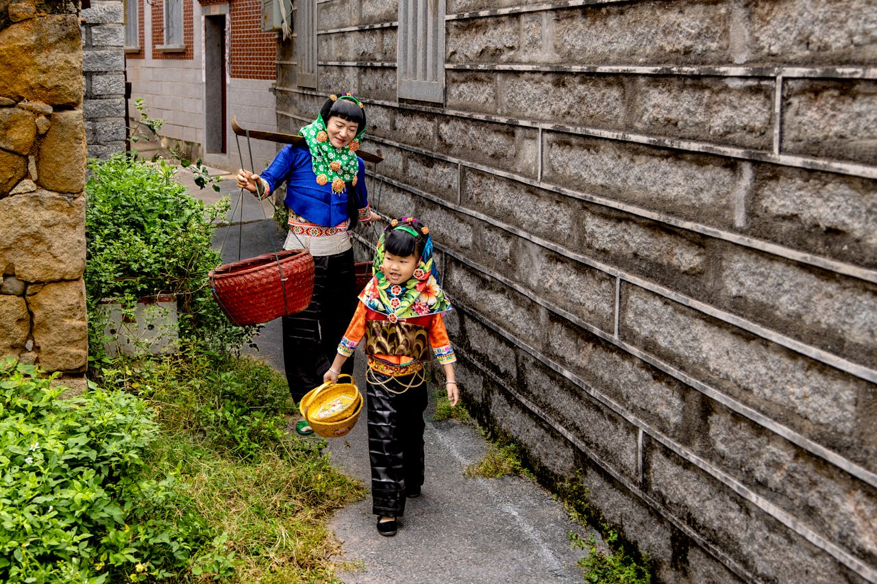gallery-2 Mother and child in traditional attire carrying baskets in a village alley.