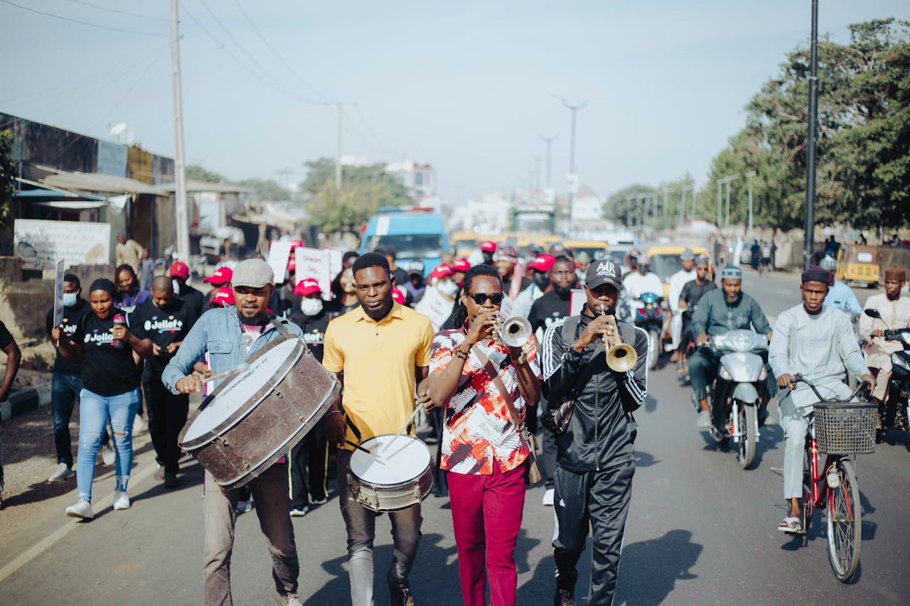 gallery-1 Vibrant street parade with musicians playing drums and trumpets, leading a diverse crowd.